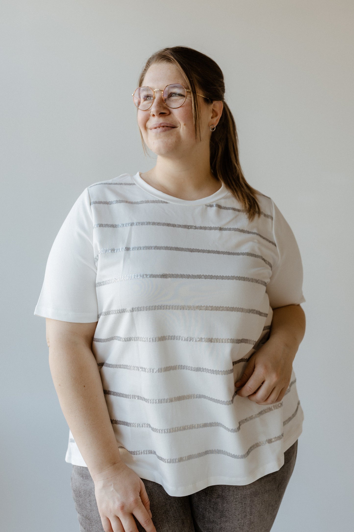 A person with glasses and a ponytail, wearing a Sequin Stripe Tee made of soft fabric and gray pants, stands against a plain background, looking slightly to the side.