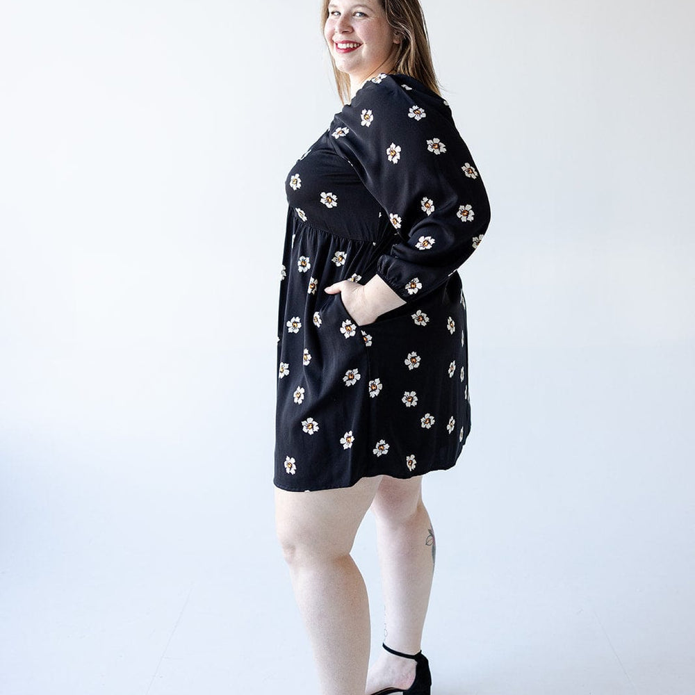 A woman models a Mikarose LONG-SLEEVED BABYDOLL KNEE LENGTH DRESS, featuring a black floral pattern, paired with black sandals as she smiles against a plain white background.