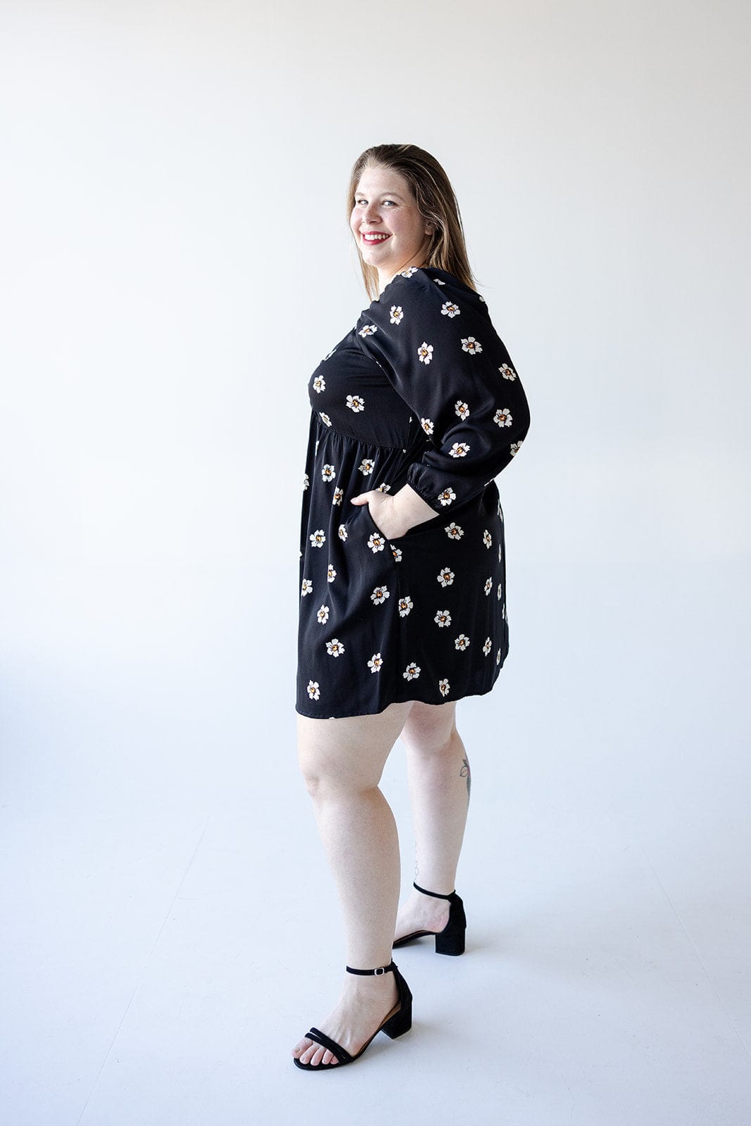 A woman models a Mikarose LONG-SLEEVED BABYDOLL KNEE LENGTH DRESS, featuring a black floral pattern, paired with black sandals as she smiles against a plain white background.