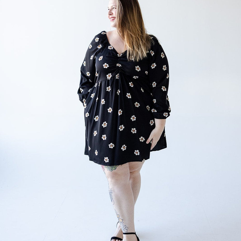 A person wearing a Mikarose long-sleeved babydoll knee-length dress, which features white floral patterns against a black background, stands in a bright, minimalist space, smiling and looking to the side.