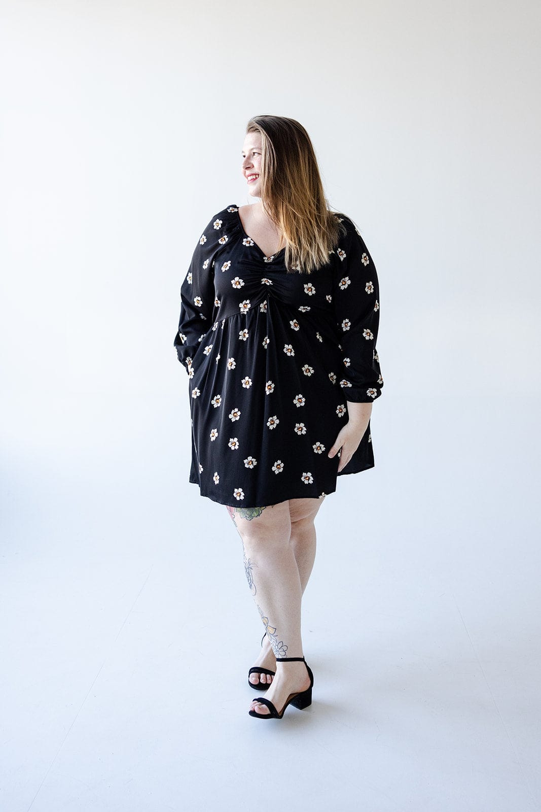 A person wearing a Mikarose long-sleeved babydoll knee-length dress, which features white floral patterns against a black background, stands in a bright, minimalist space, smiling and looking to the side.