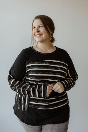 A person with long hair is smiling and looking to the side, wearing a black-and-white striped shirt with subtle stripe detail and gray pants, standing against a plain background.