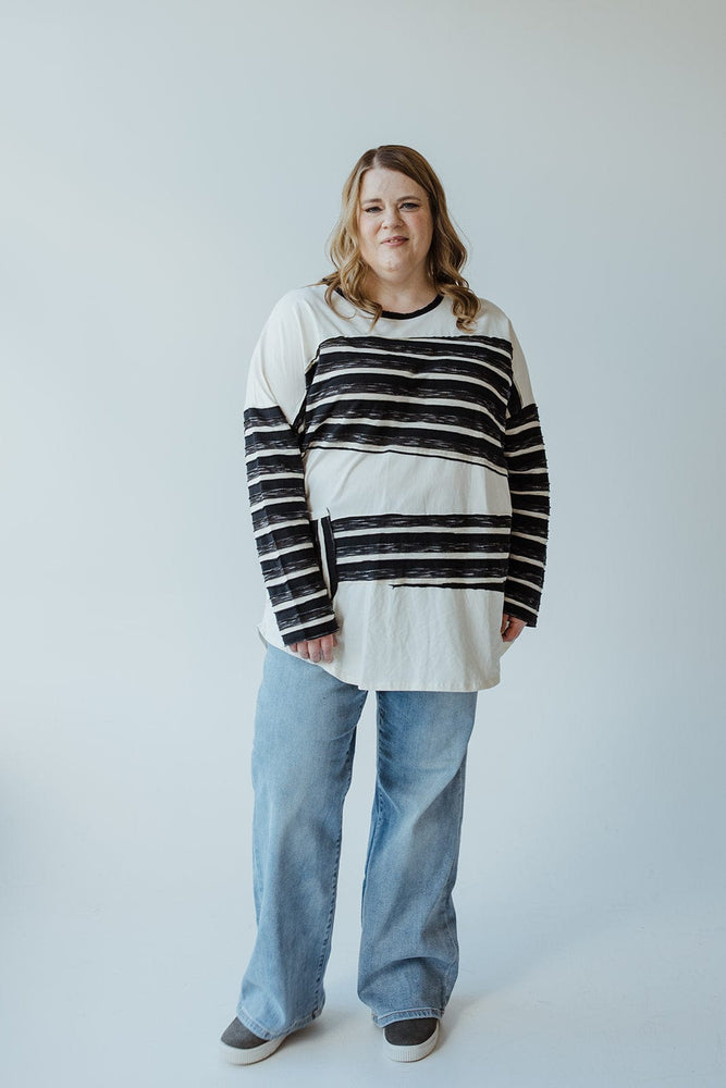 A person with long hair stands facing the camera, wearing a mid-size LONG-SLEEVE ROUND NECK TUNIC WITH STRIPE DETAIL IN CREAM by Love Marlow, light blue jeans, and gray shoes. They are holding a folded tablet in one hand.