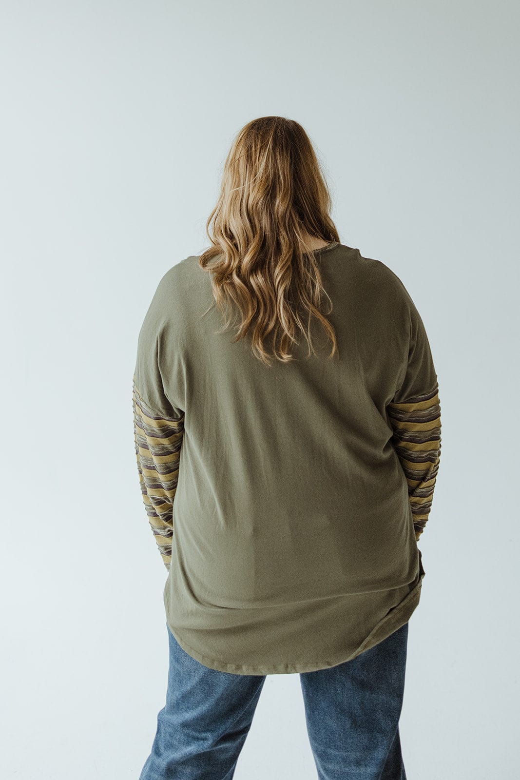 A person with luxurious long hair is seen from the back, wearing a neon buddha LONG-SLEEVE ROUND NECK TUNIC WITH STRIPE DETAIL IN KALE and jeans that create a modern profile.