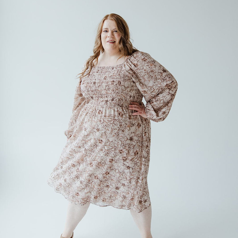 A person with long hair stands confidently in a Love Marlow LONG BLOUSON SLEEVED SMOCKED BODICE DRESS IN SIMPLY ROSEY BEIGE and sandals, the rosey beige color of their outfit contrasting beautifully against the plain white background.