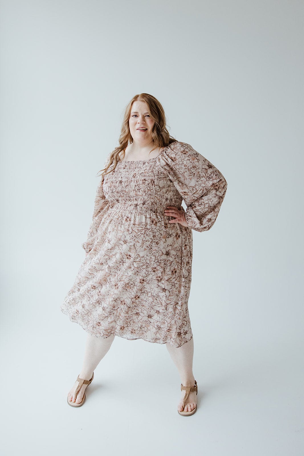 A person with long hair stands confidently in a Love Marlow LONG BLOUSON SLEEVED SMOCKED BODICE DRESS IN SIMPLY ROSEY BEIGE and sandals, the rosey beige color of their outfit contrasting beautifully against the plain white background.