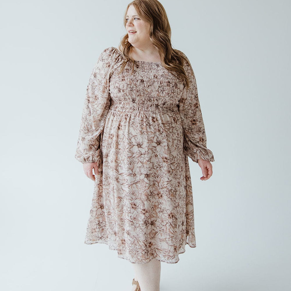 A woman wearing a LONG BLOUSON SLEEVED SMOCKED BODICE DRESS IN SIMPLY ROSEY BEIGE by Love Marlow with sandals stands against a plain white background, looking to her left.