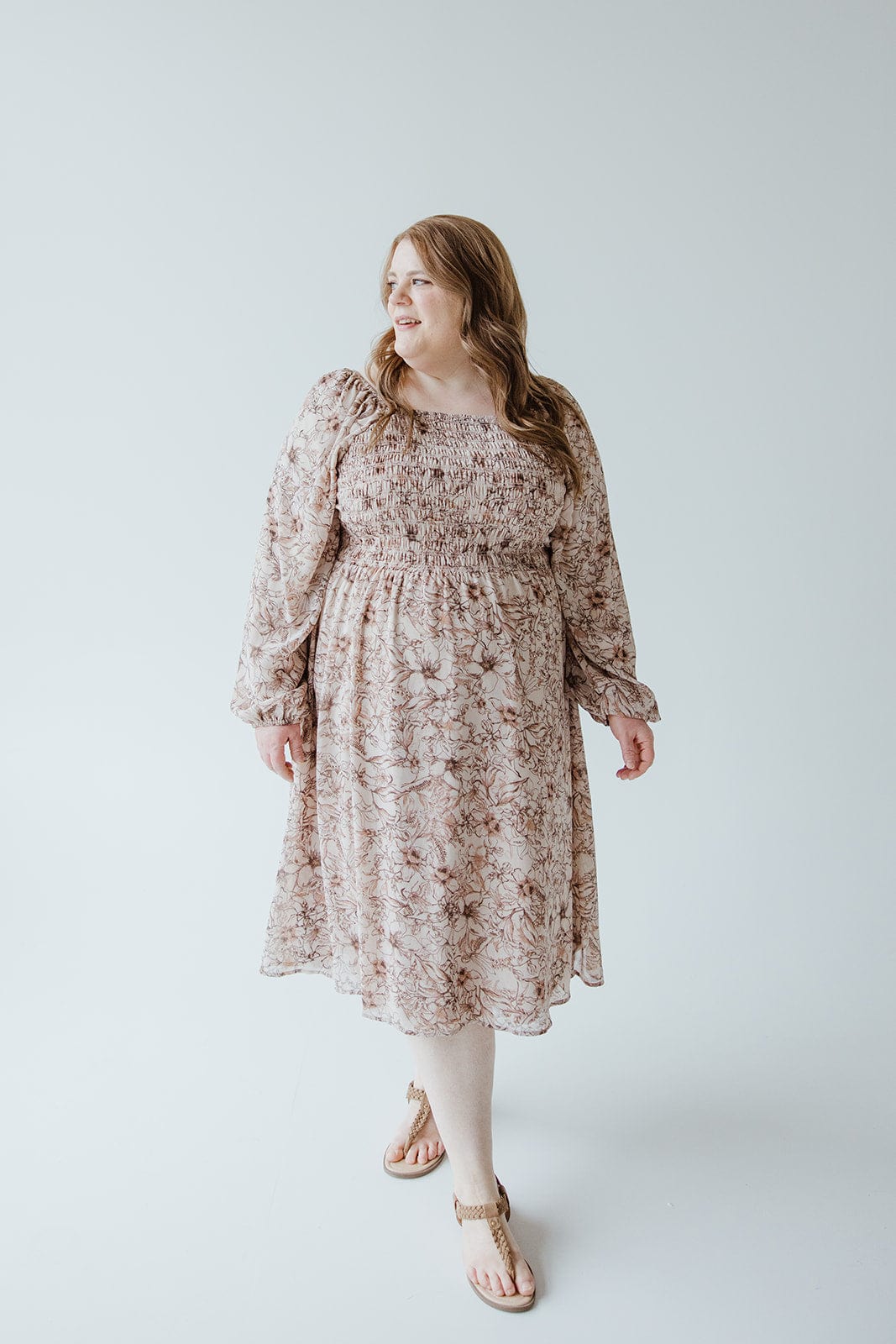 A woman wearing a LONG BLOUSON SLEEVED SMOCKED BODICE DRESS IN SIMPLY ROSEY BEIGE by Love Marlow with sandals stands against a plain white background, looking to her left.