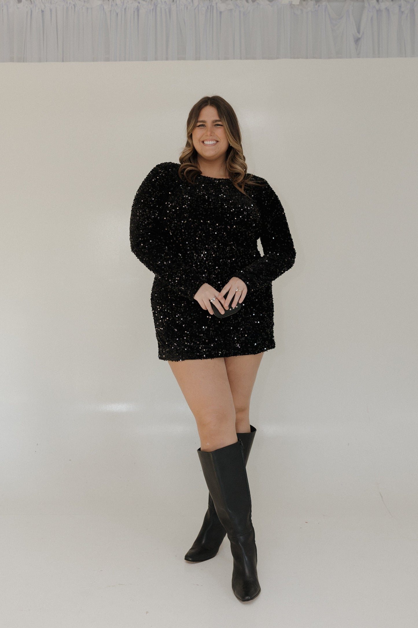 Woman wearing a black sparkly dress and knee-high boots against a white background