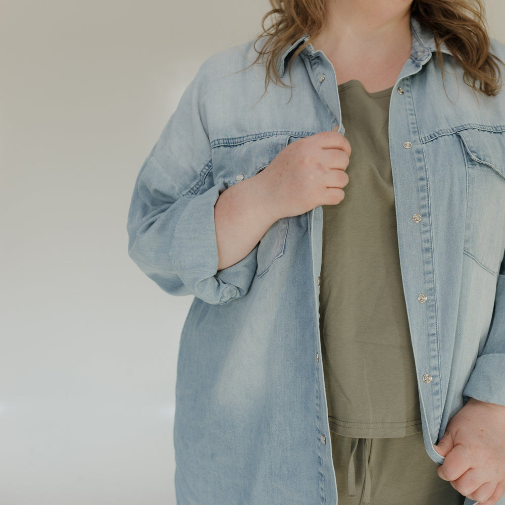Person wearing a light blue denim jacket over a green shirt and beige pants against a plain background