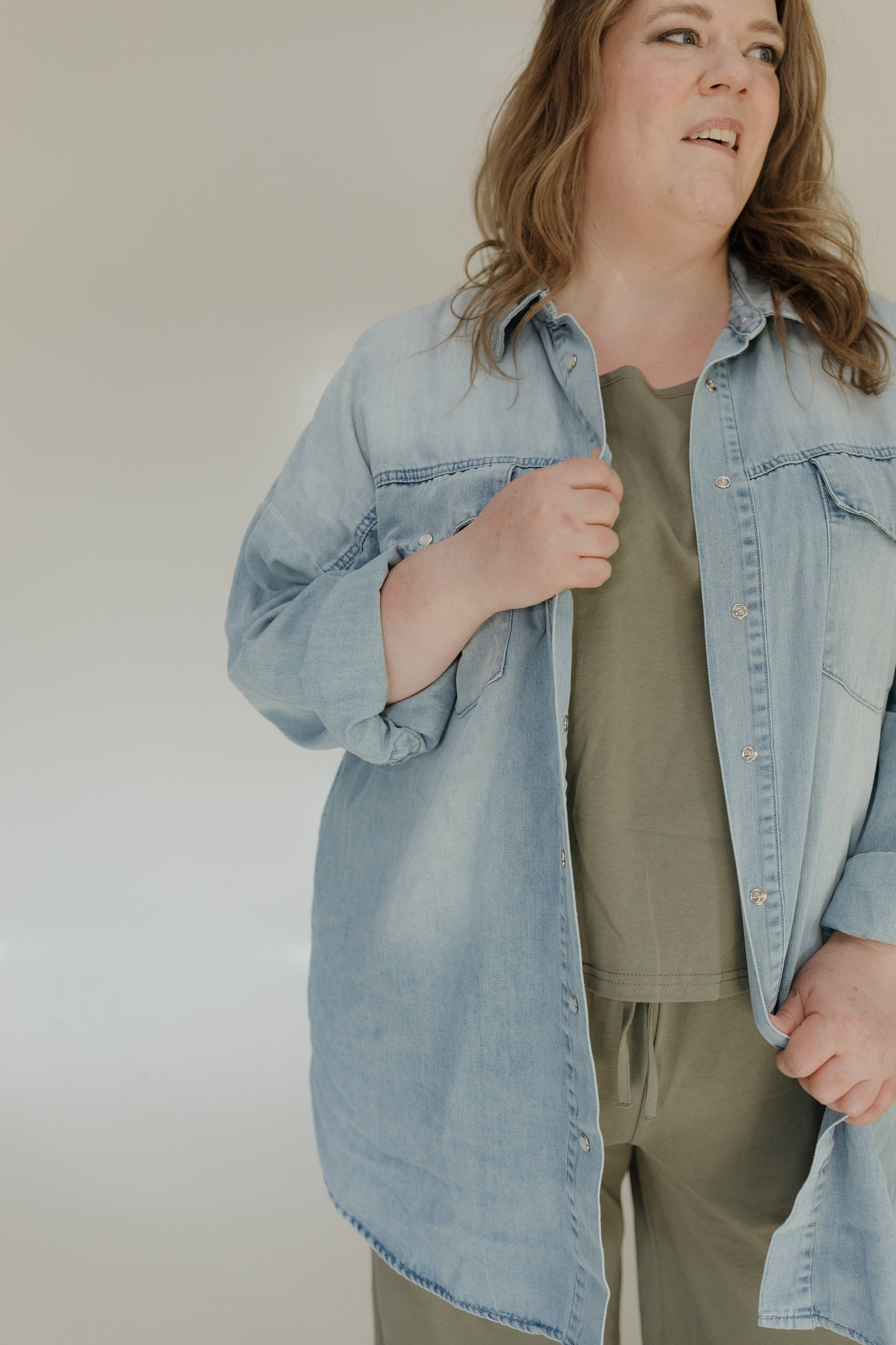 Person wearing a light blue denim jacket over a green shirt and beige pants against a plain background