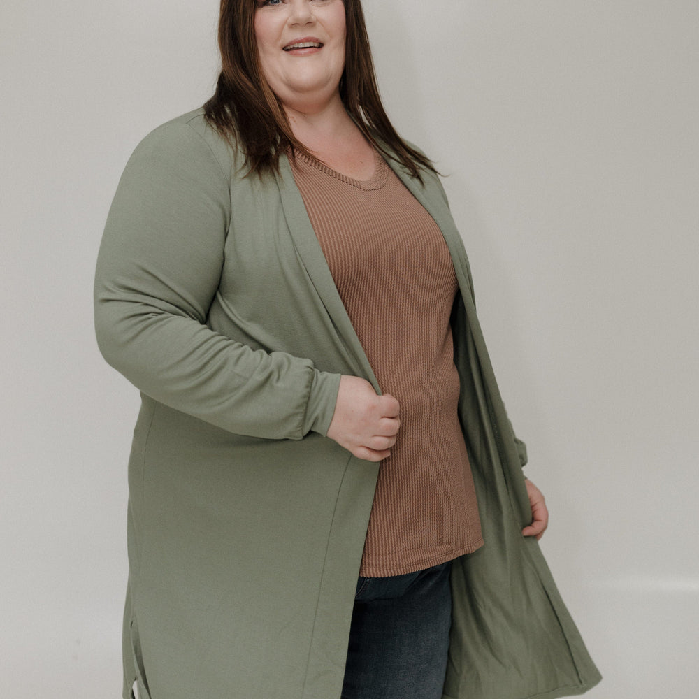 Woman wearing a green cardigan over a brown shirt and blue jeans on a plain background