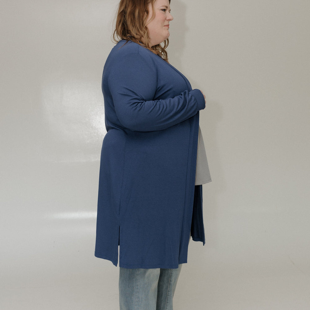 Person wearing a blue coat and jeans standing against a white wall.