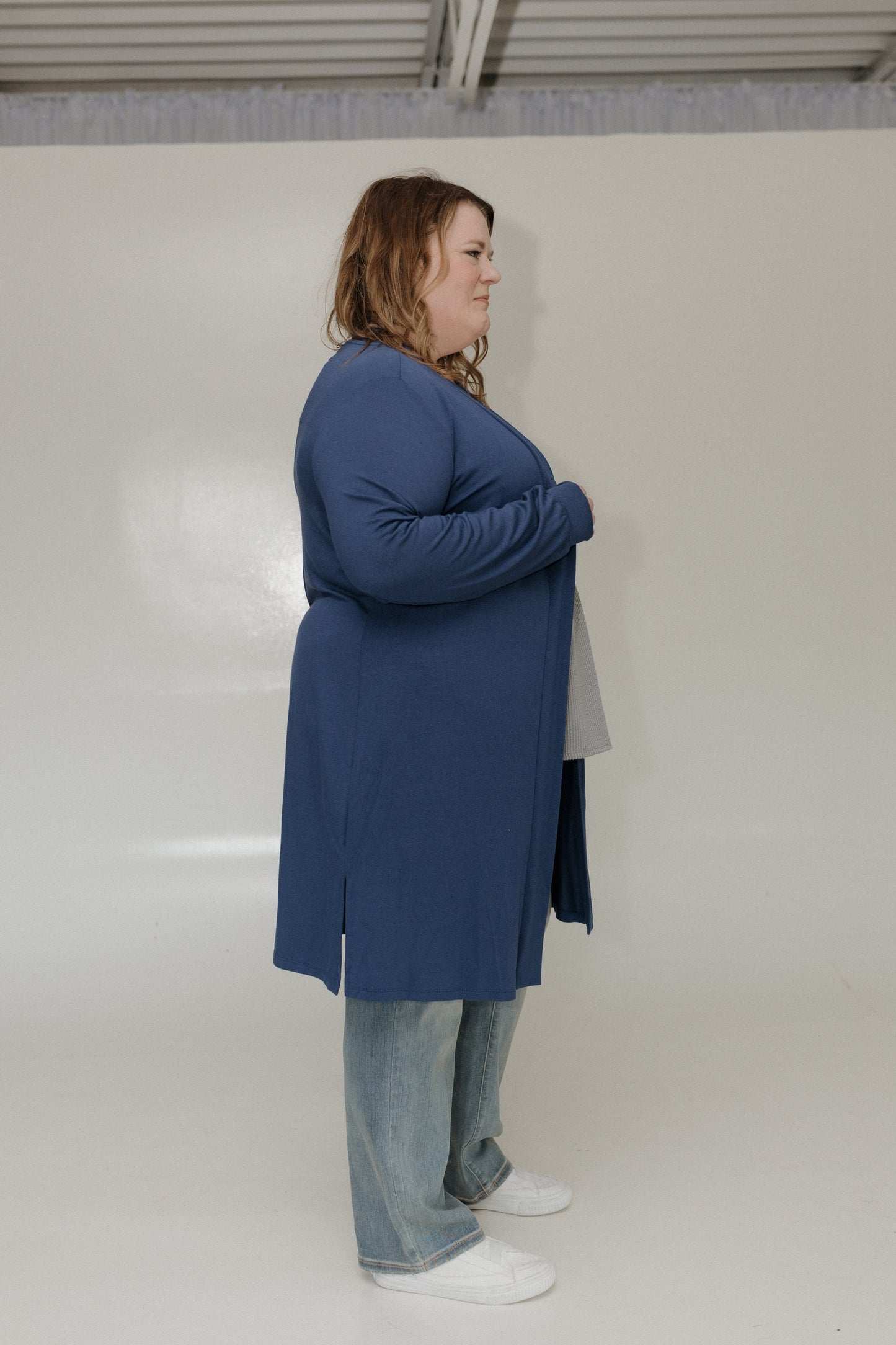 Person wearing a blue coat and jeans standing against a white wall.