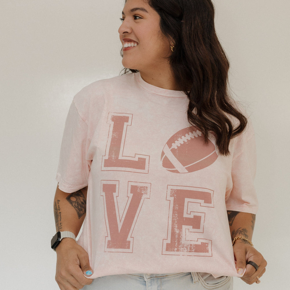 "LOVE" FOOTBALL GRAPHIC TEE IN LIGHT BLUSH MINERAL WASH Woman wearing a pink 'LOVE' t-shirt with a football graphic on a plain A person stands smiling in a Love Marlow "LOVE" FOOTBALL GRAPHIC TEE IN LIGHT BLUSH MINERAL WASH, paired with blue pants and flip-flops, posing against a plain background.