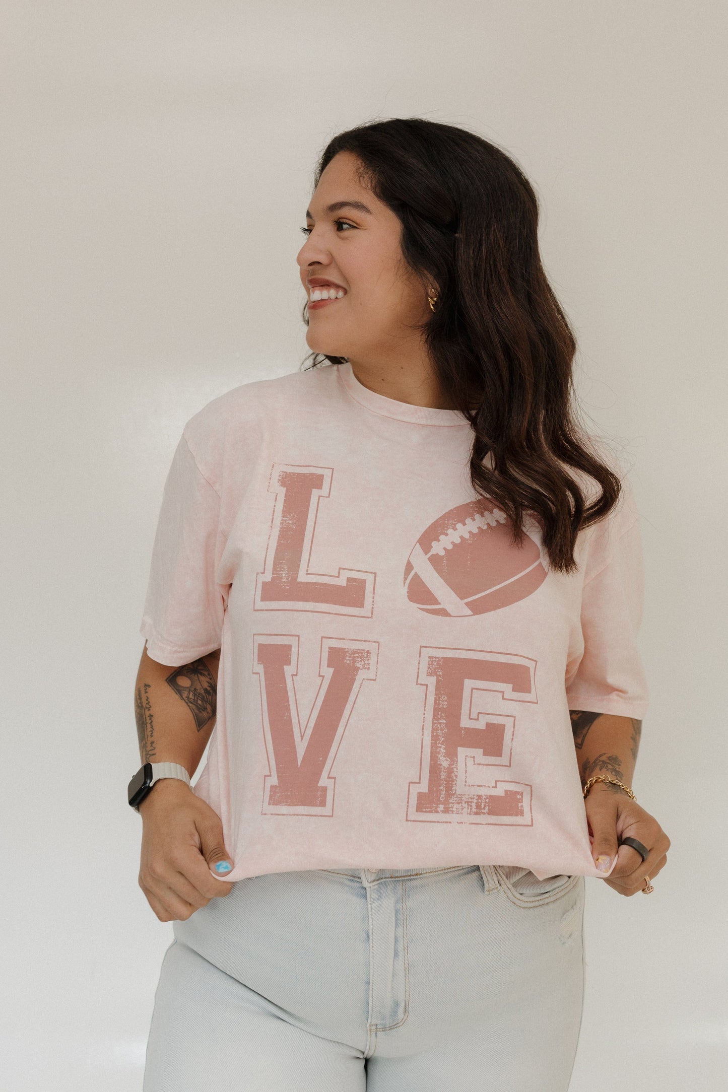 "LOVE" FOOTBALL GRAPHIC TEE IN LIGHT BLUSH MINERAL WASH Woman wearing a pink 'LOVE' t-shirt with a football graphic on a plain A person stands smiling in a Love Marlow "LOVE" FOOTBALL GRAPHIC TEE IN LIGHT BLUSH MINERAL WASH, paired with blue pants and flip-flops, posing against a plain background.