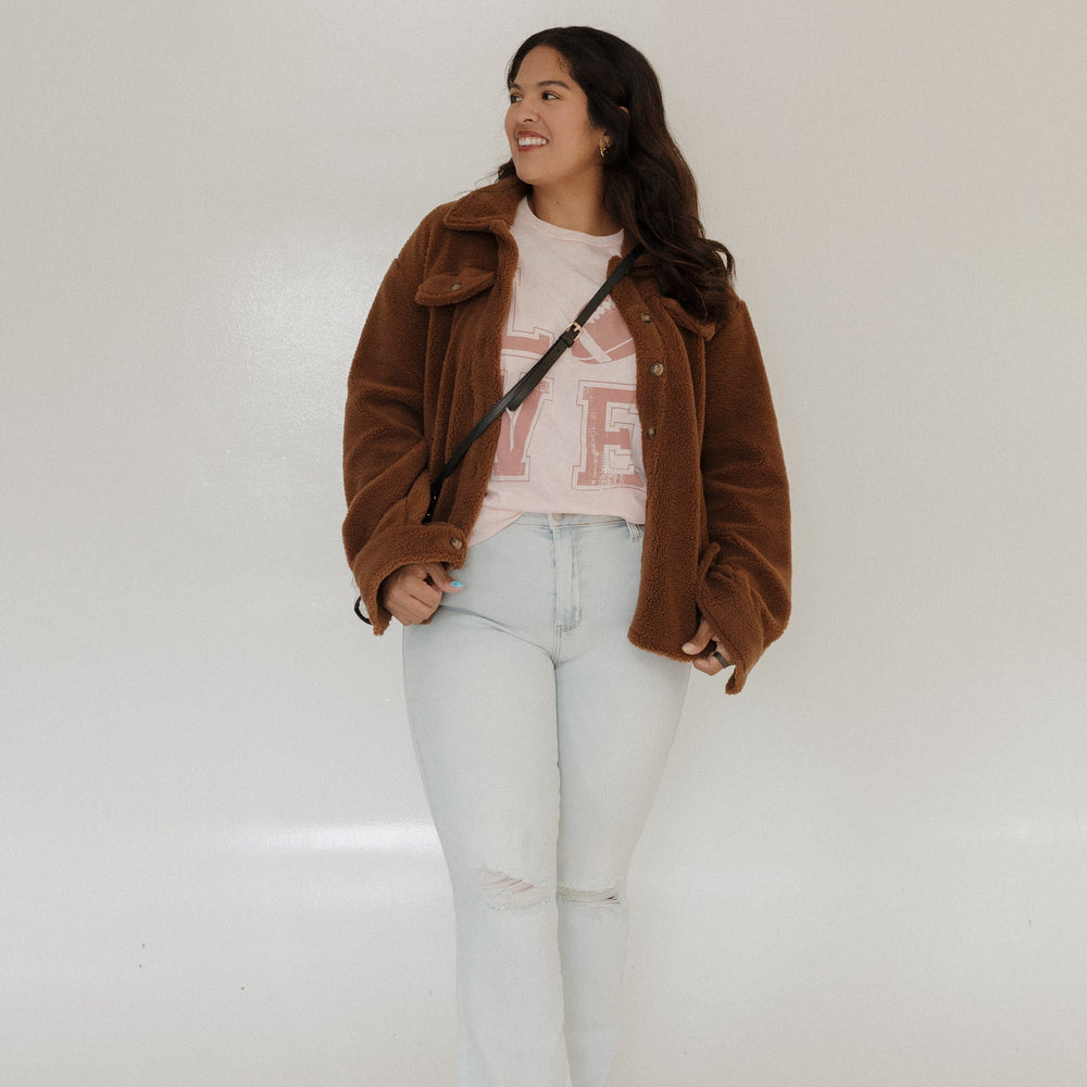 "LOVE" FOOTBALL GRAPHIC TEE IN LIGHT BLUSH MINERAL WASH Woman wearing a pink 'LOVE' t-shirt with a football graphic on a plain A person stands smiling in a Love Marlow "LOVE" FOOTBALL GRAPHIC TEE IN LIGHT BLUSH MINERAL WASH, paired with blue pants and flip-flops, posing against a plain background.