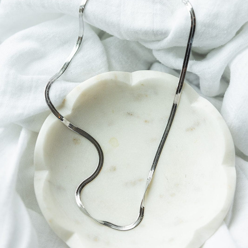 The LUXE SILVER DELICATE HERRINGBONE CHAIN by Love Marlow, featuring a sleek and twisted design in luxurious silver, elegantly rests on a white fabric background and a round dish. This versatile necklace is crafted from hypoallergenic stainless steel.