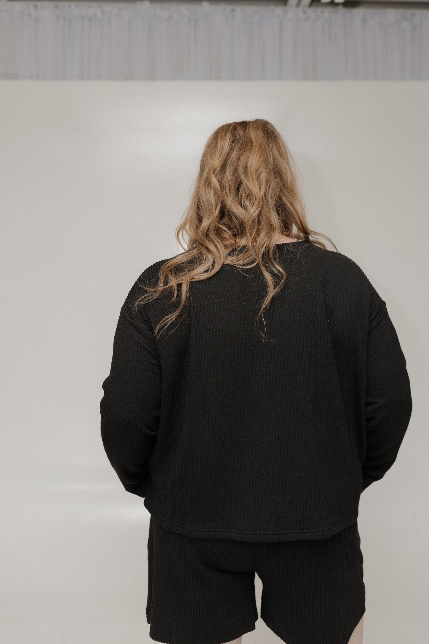 Person wearing a black outfit with long hair, standing against a plain background