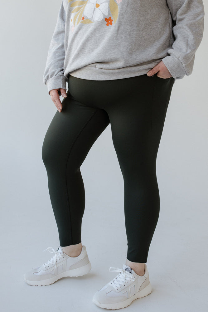 A person wearing a gray sweatshirt, Spanx® SPANXshape™ Booty Boost® Active 7/8 Leggings in Essex Green, and white sneakers stands with one hand in a pocket on a light background.