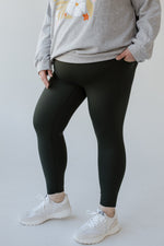 A person wearing a gray sweatshirt, Spanx® SPANXshape™ Booty Boost® Active 7/8 Leggings in Essex Green, and white sneakers stands against a plain background.