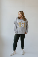 A person wearing a gray floral sweater, Spanx® SPANXshape™ Booty Boost® Active 7/8 Leggings, and white sneakers stands against an Essex Green background.