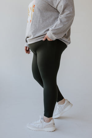 A person in sculpting performance fabric, wearing Essex Green Spanx SPANXshape Booty Boost Active 7/8 Leggings and a grey shirt, stands poised.