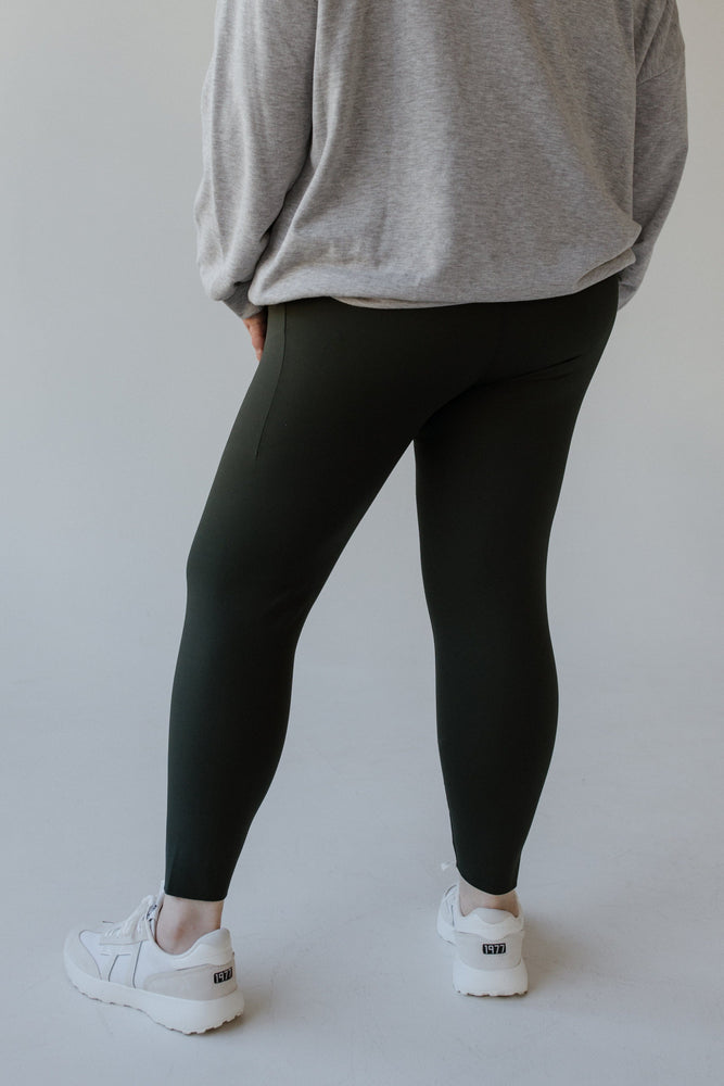 A person wearing a gray sweatshirt, Spanx® SPANXshape™ Booty Boost® Active 7/8 Leggings in Essex Green, and white sneakers stands against a plain background.