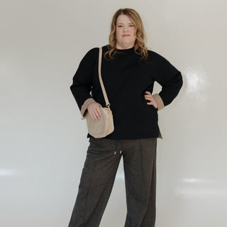 Woman wearing a black sweater and gray pants with a beige handbag against a white background