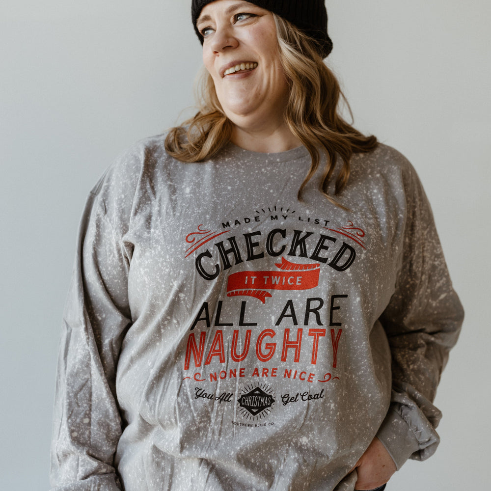 A person in a beanie and gray hand-bleached sweater, featuring the funny graphic text "Checked it twice, all are naughty, none are nice," stands smiling against a plain background, exuding holiday spirit.