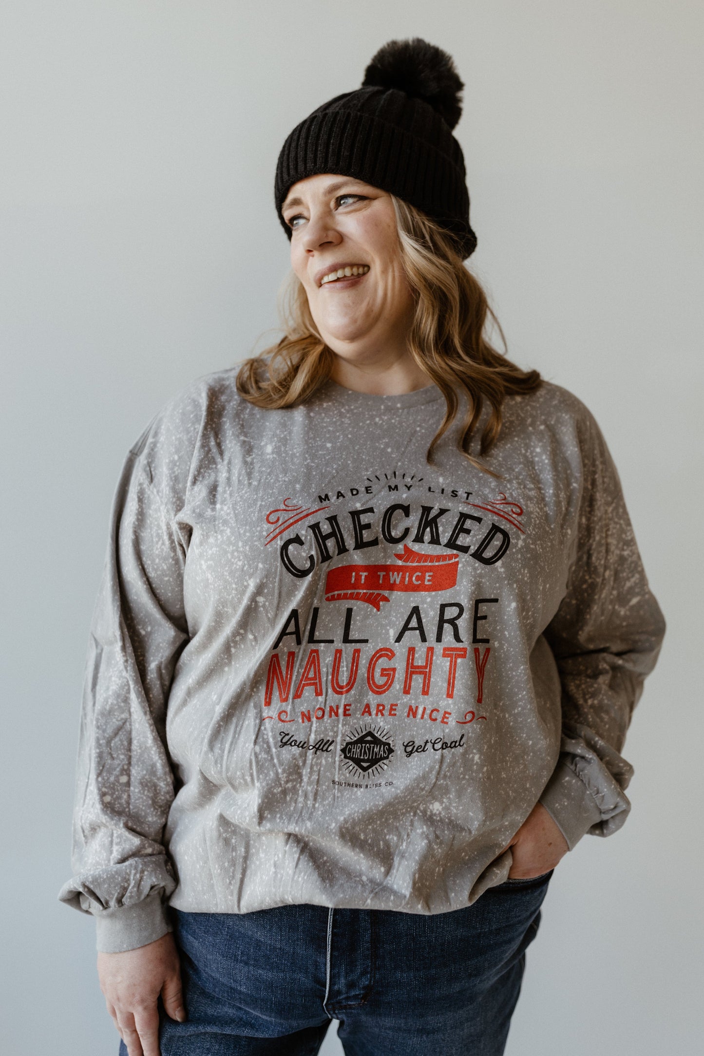 A person in a beanie and gray hand-bleached sweater, featuring the funny graphic text "Checked it twice, all are naughty, none are nice," stands smiling against a plain background, exuding holiday spirit.