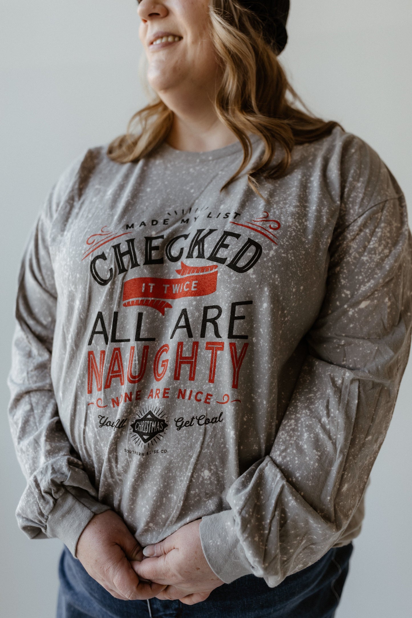 Person wearing a gray, hand-bleached sweater with the text: "Made the list, checked it twice, all are naughty, none are nice. Christmas get real." A funny graphic tee that captures the holiday spirit perfectly.