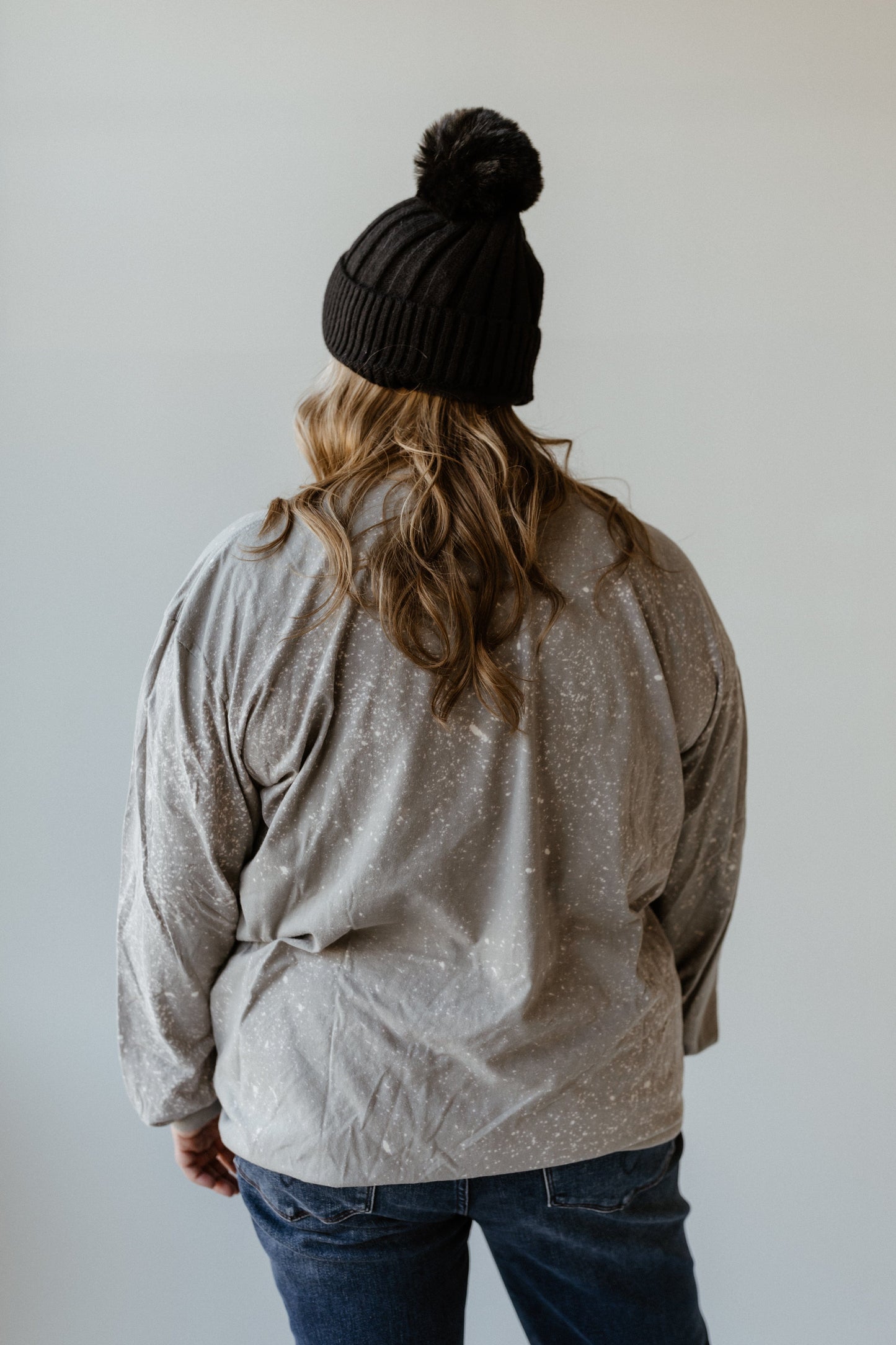 A person with long hair in a black beanie and a hand bleached, speckled gray sweater faces away from the camera.