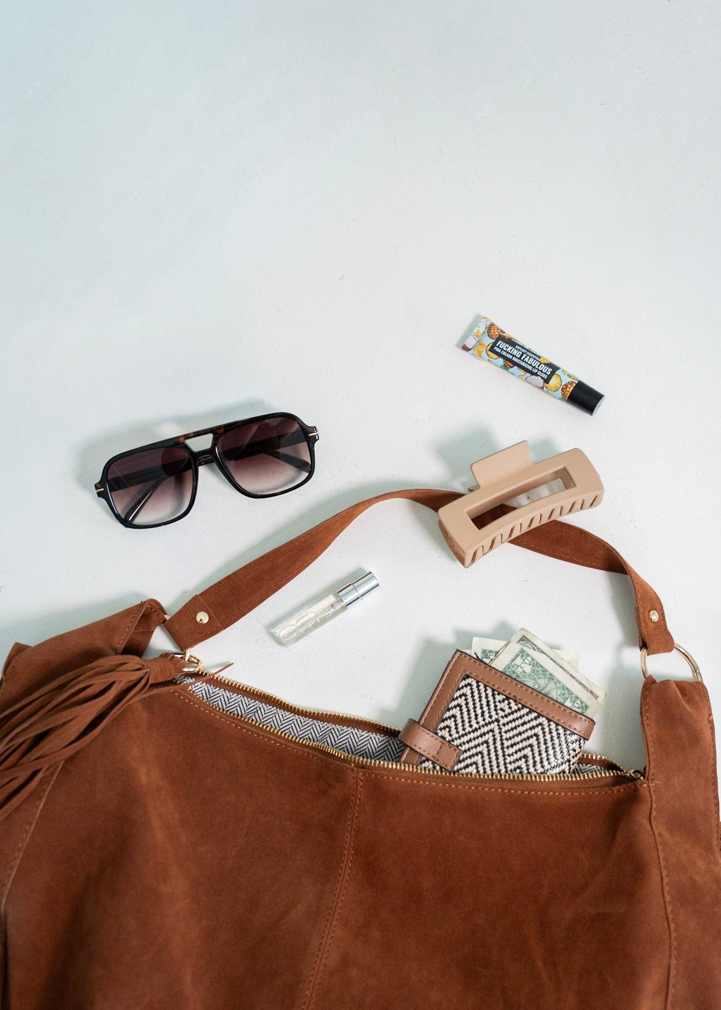 Brown leather bag with sunglasses, hair clip, and money on a white background