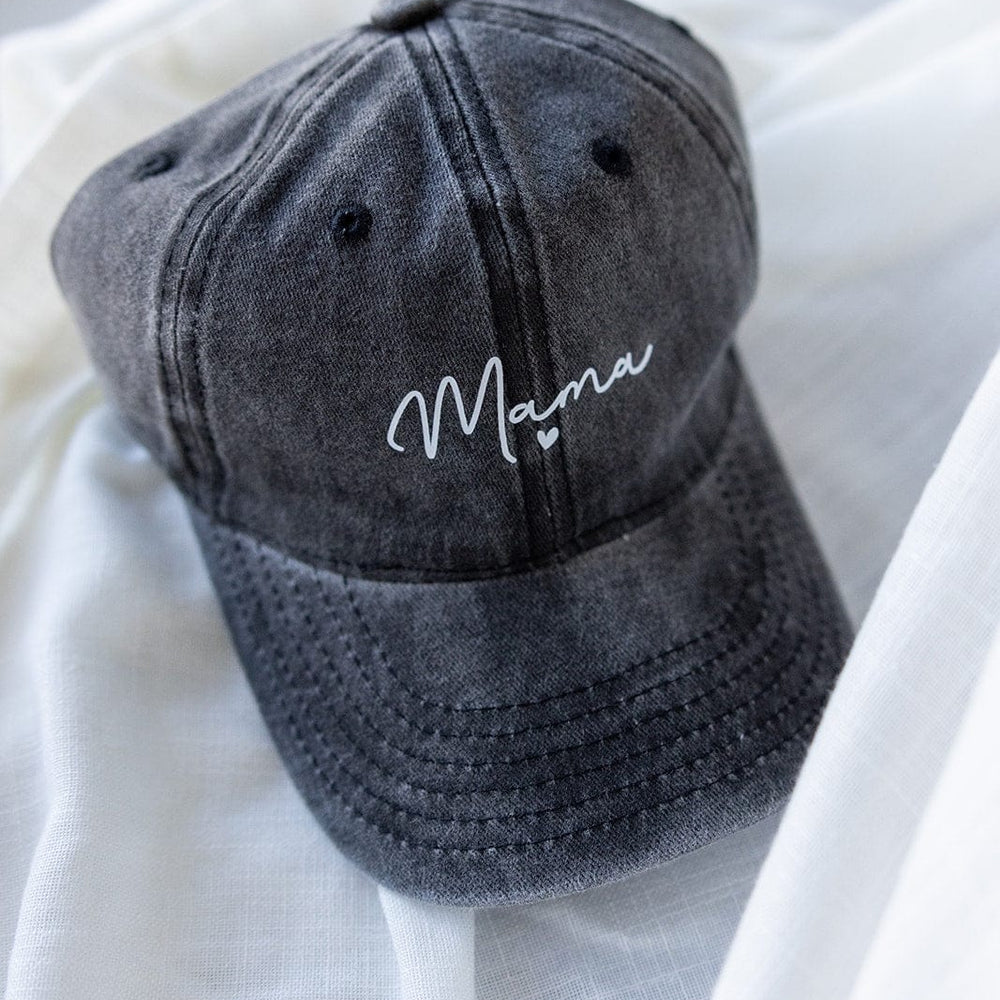 A dark gray, acid-washed baseball cap with the word “Mama” in white cursive lettering is elegantly placed on white fabric, making it a heartfelt choice for Mother's Day gifting.