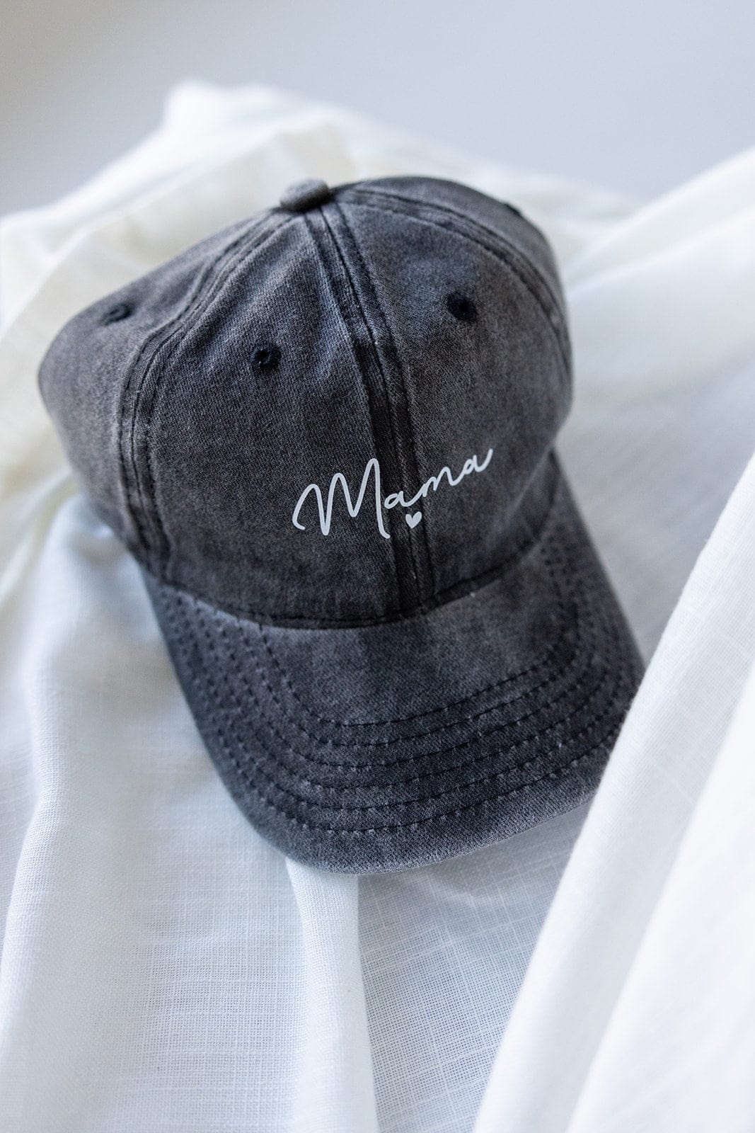 A dark gray, acid-washed baseball cap with the word “Mama” in white cursive lettering is elegantly placed on white fabric, making it a heartfelt choice for Mother's Day gifting.