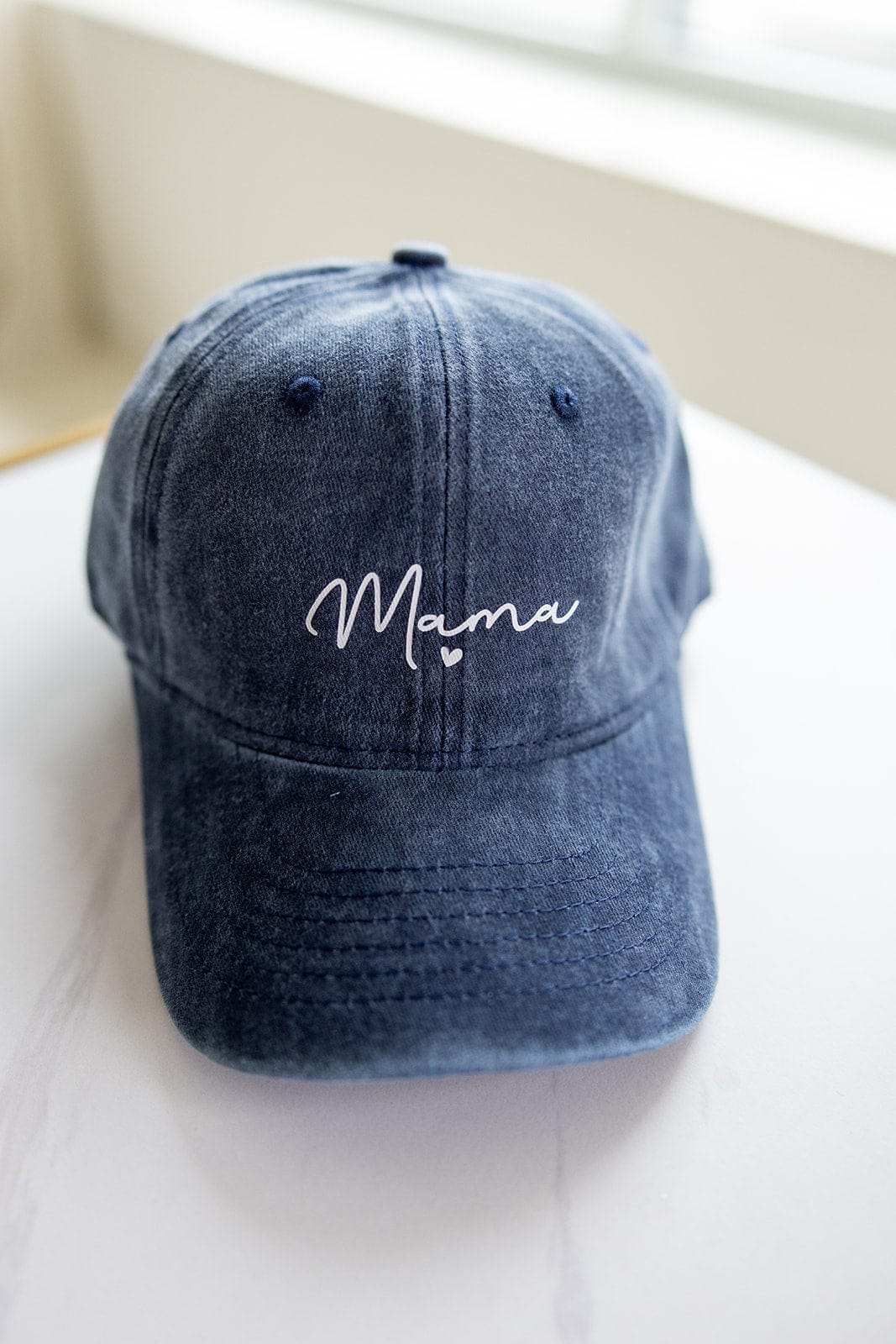 The "MAMA" Acid Washed Baseball Cap in Navy by Love Marlow, with "Mama" elegantly embroidered in white on the front, sits stylishly on a light surface—making it the perfect Mother's Day gift to add a special touch of love.