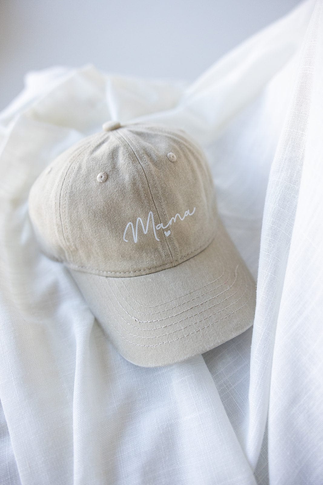 Beige MAMA Baseball Cap with "Mama" embroidered in white on the front, featuring an acid-washed design. Perfectly displayed on a soft white fabric, making it an ideal Mother's Day gift.