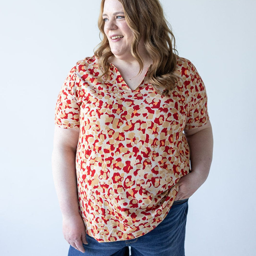 Person with shoulder-length wavy hair, wearing a red and beige patterned MANDARIN COLLAR JERSEY KNIT BLOUSE by Love Marlow and denim shorts, stands against a plain white background, looking off to the side and smiling.