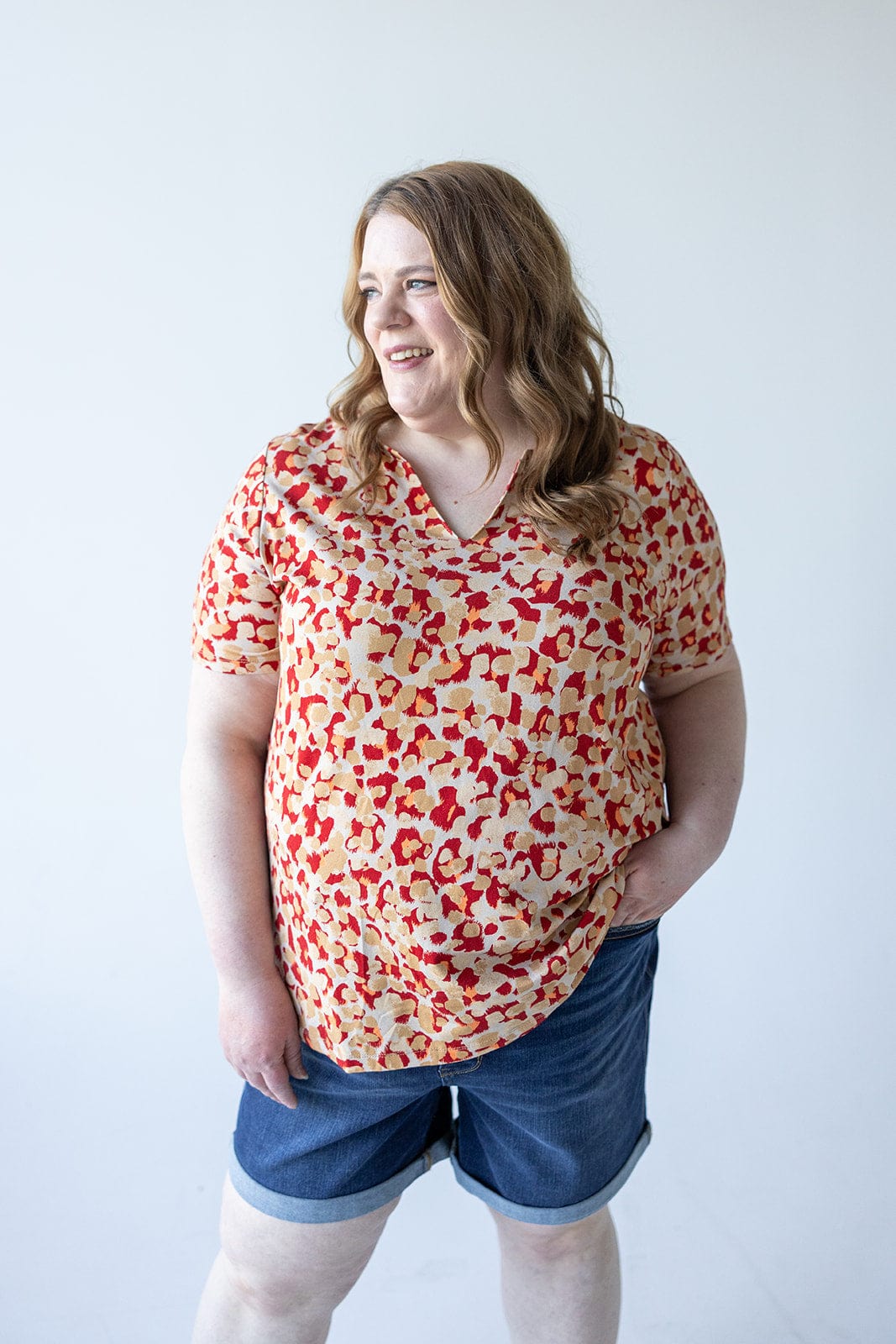 Person with shoulder-length wavy hair, wearing a red and beige patterned MANDARIN COLLAR JERSEY KNIT BLOUSE by Love Marlow and denim shorts, stands against a plain white background, looking off to the side and smiling.