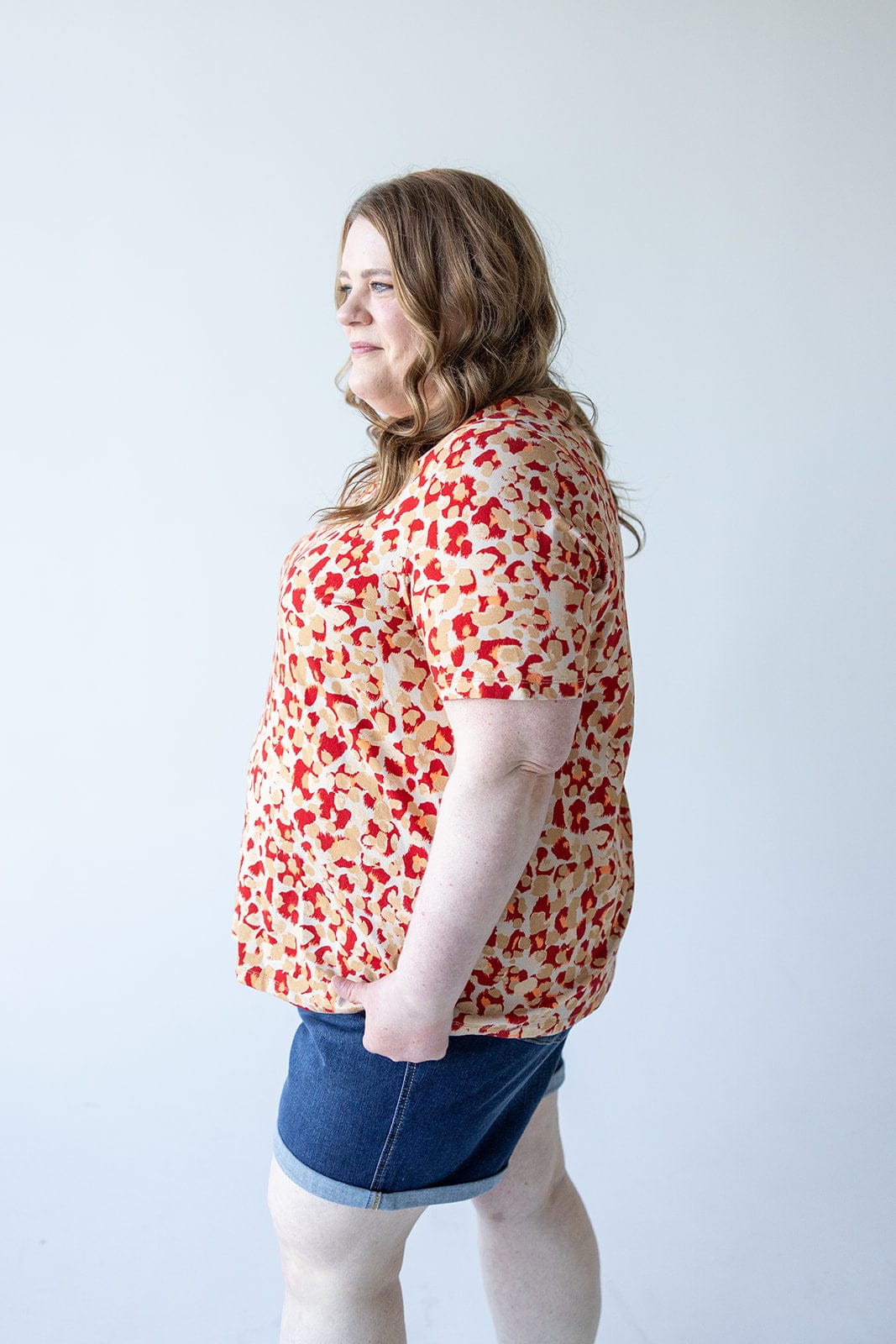 A person with long, wavy hair stands in profile wearing a MANDARIN COLLAR JERSEY KNIT BLOUSE from Love Marlow and denim shorts made of jersey knit fabric against a plain background.