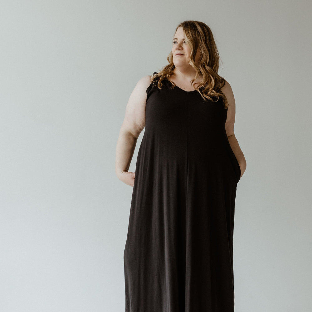 A person with long hair wearing a black sleeveless slip dress stands against a plain background, looking to the side with hands in pockets. The dress features adjustable cami straps, offering both style and comfort.