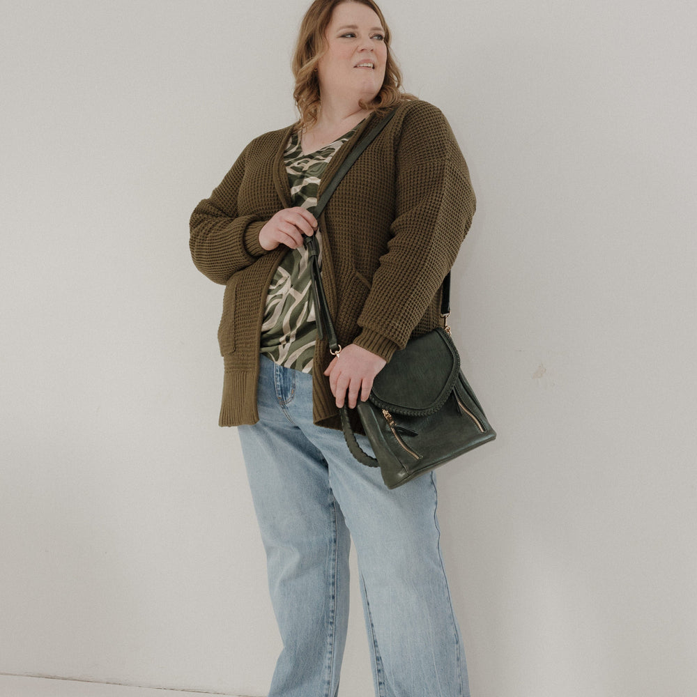 Woman wearing a brown cardigan, patterned top, and jeans, holding a green leather bag against a white background