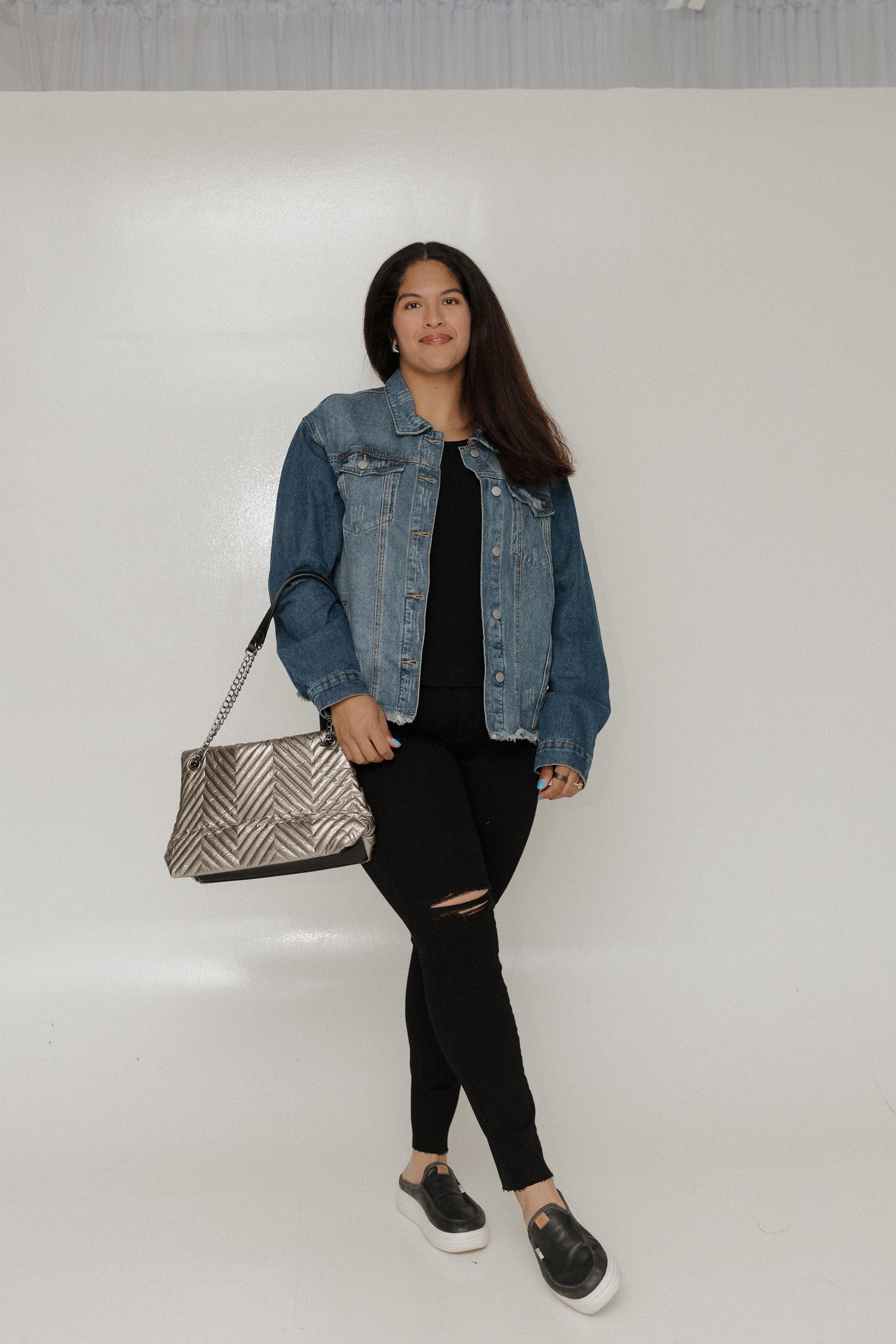 Woman wearing a denim jacket, black outfit, and holding a patterned handbag on a white background