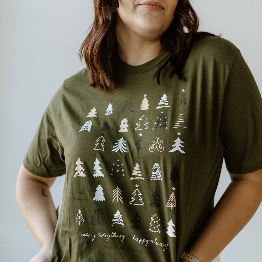 A person embraces the holiday spirit in a cozy style, sporting a green Christmas tee adorned with a tree pattern and the cheerful message "merry everything & happy always.