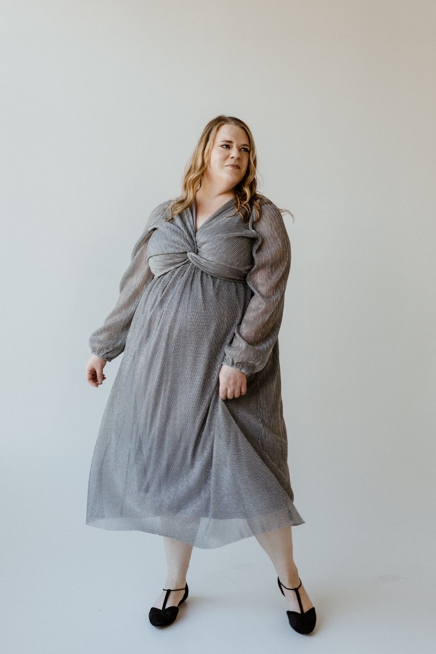 A plus-size fashionista gracefully poses in a long, metallic silver plisse dress against a plain backdrop.