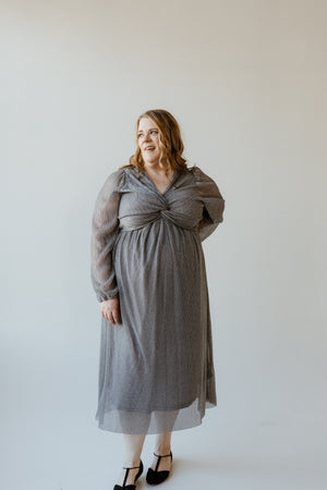 A plus-size fashionista gracefully stands against a plain white background, wearing a long, textured metallic silver plisse dress with black shoes and a slight smile, subtly angled to the side.