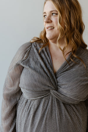 A plus-size fashionista with long wavy hair smiles, wearing a shimmery metallic silver plisse dress with a twisted front design.