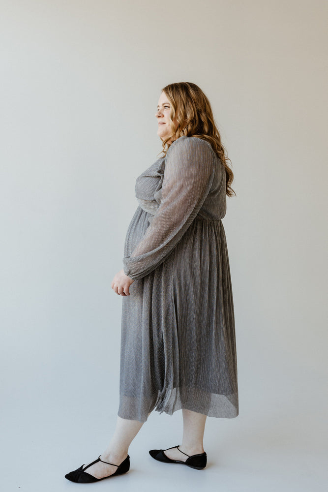 A plus-size fashionista stands gracefully against a light background, wearing a flowing metallic silver plisse dress and black shoes, her gaze directed to the left.