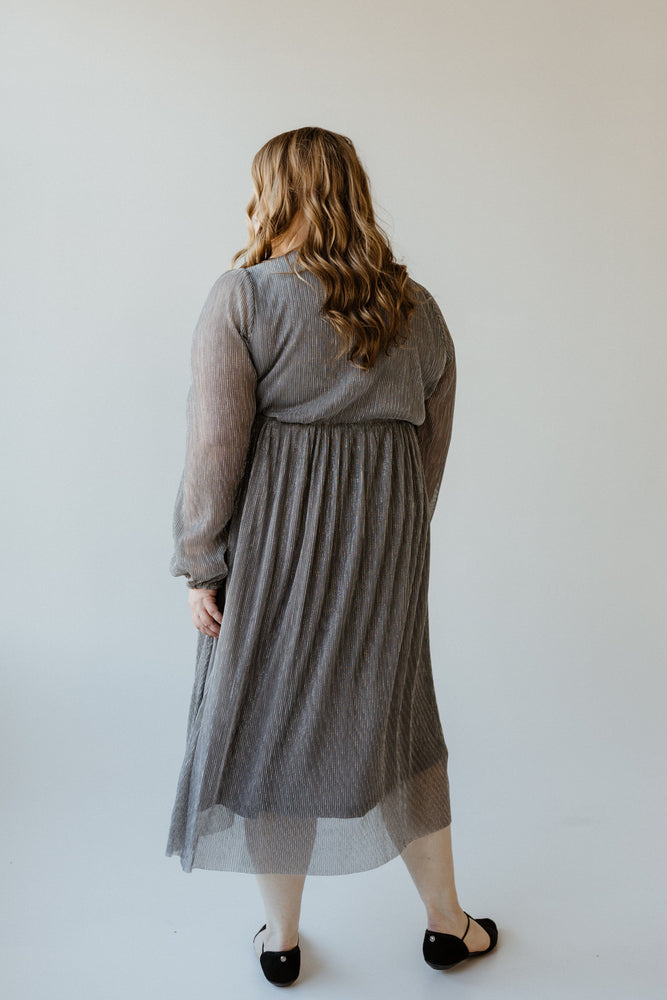 A plus-size fashionista with long hair stands facing away, wearing a metallic silver plisse dress and black shoes, against a plain background.